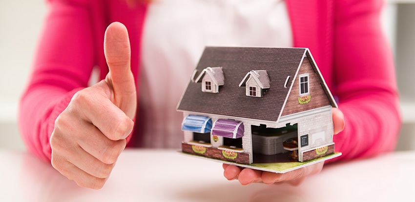 photo of a mortgage broker holding up a small model home and giving a thumbs up, showing that it helps to hire a mortgage broker to get approved for a home loan